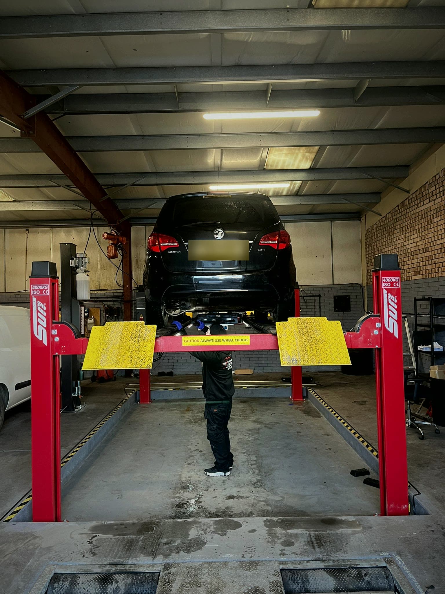A car raised on the red four-post lift in our Unit 2 MOT centre, a technician underneath.
